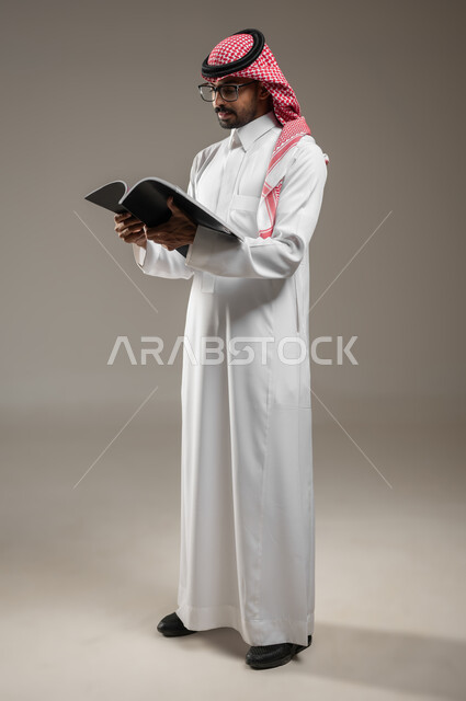 قضاء الوقت بالقراءة والتعلم والثقافة، التمعن والتركيز، بورتريه لرجل عربي سعودي خليجي يرتدي الزي السعودي التقليدي يحمل بيده كتاب، ينظر الى الكتاب بتركيز وتمعن، صوره الجسم بالطول الكامل، خلفية رماديه، محتوى حساس، استخدام حساس