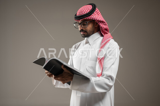 Spending time reading, learning and culture, contemplating and concentrating, a close-up portrait of a Saudi Gulf Arab man wearing traditional Saudi clothing holding a book in his hand, looking at the book with concentration and contemplation, gray background, sensitive content, sensitive use