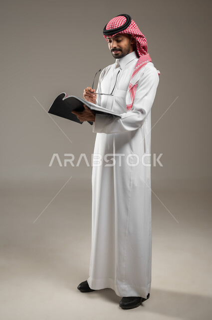 قضاء الوقت بالقراءة والتعلم والثقافة، التمعن والتركيز، بورتريه لرجل عربي سعودي خليجي يرتدي الزي السعودي التقليدي يحمل بيده كتاب، ينظر الى الكتاب بتركيز وتمعن، صوره الجسم بالطول الكامل، خلفية رماديه، محتوى حساس، استخدام حساس