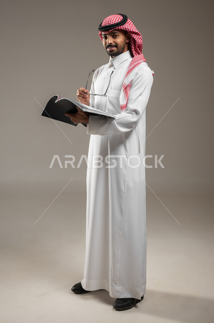 قضاء الوقت بالقراءة والتعلم والثقافة، التمعن والتركيز، بورتريه لرجل عربي سعودي خليجي يرتدي الزي السعودي التقليدي يحمل بيده كتاب، ينظر الى الكاميرا بابتسامه، صوره الجسم بالطول الكامل، خلفية رماديه، محتوى حساس، استخدام حساس