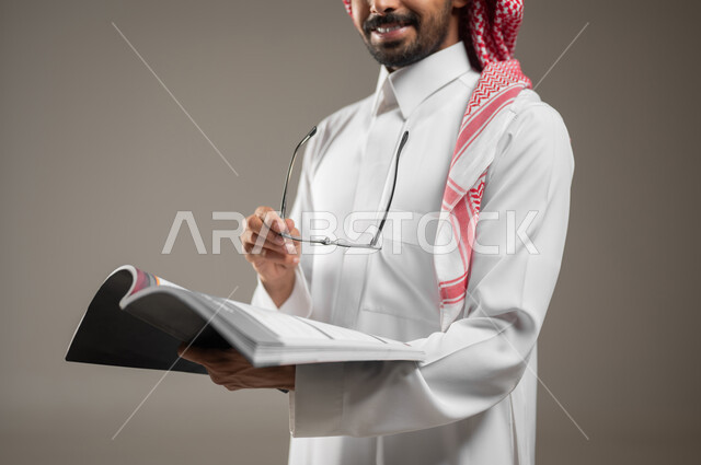 قضاء الوقت بالقراءة والتعلم والثقافة، التمعن والتركيز، بورتريه مقرب لرجل عربي سعودي خليجي يرتدي الزي السعودي التقليدي يحمل بيده كتاب، ينظر الى الكاميرا بابتسامه، خلفية رماديه، محتوى حساس، استخدام حساس