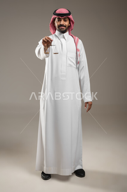 بورتريه لرجل عربي خليجي سعودي يرتدي الزي السعودي، يحمل بيده ميزان العدل ، العمل في مجال القضاء والمحاماة ، القانون والمحاماة ، دراسة القانون، مفهوم العدالة والقانون ، مهن و وظائف قانونية ، صوره الجسم بالطول الكامل، خلفية رماديه، استخدام حساس، محتوى حساس