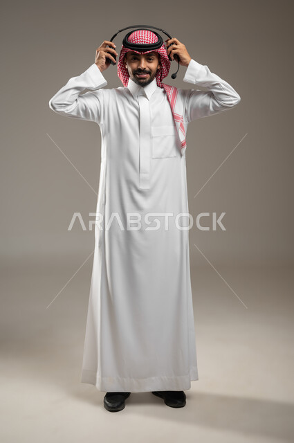 Using headphone technology, responding to customers and inquiries, a portrait of a Saudi Gulf Arab man wearing an audio headset holding it in his hand, electronic marketing from home, Saudi administrative professions and jobs, Saudi Gulf employees, full-length body photo, gray background, sensitive content