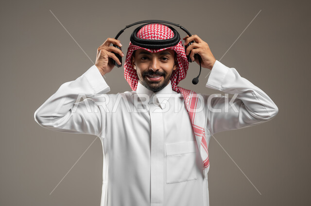 Using headphone technology, responding to customers and inquiries, a close-up portrait of a Saudi Gulf Arab man wearing an audio headset holding it in his hand, electronic marketing from home, Saudi administrative professions and jobs, Saudi Gulf employees, gray background, sensitive content, sensitive use