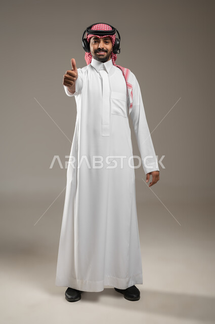 التواصل مع الأصدقاء والأقارب، استخدام تقنية الهيدفون، بورتريه لرجل عربي سعودي خليجي يرتدي الزي السعودي ويضع سماعه الرأس الصوتية يشير باصبع الابهام، ايماءات السعاده والسرور، صوره الجسم بالطول الكامل، خلفيه رماديه، استخدام حساس، محتوى حساس
