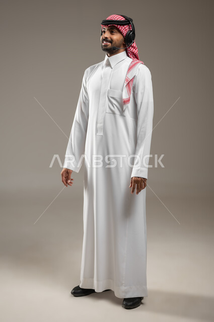 التواصل مع الأصدقاء والأقارب، استخدام تقنية الهيدفون، بورتريه لرجل عربي سعودي خليجي يرتدي الزي السعودي ويضع سماعه الرأس الصوتية ينظر الى الكاميرا بابتسامه، ايماءات السعاده والسرور، صوره الجسم بالطول الكامل، خلفيه رماديه، استخدام حساس، محتوى حساس