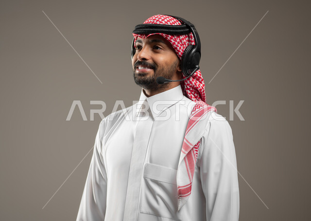 Communicating with friends and relatives, using headphone technology, a close-up portrait of a Saudi Gulf Arab man wearing Saudi clothing and wearing an audio headset, looking at the camera with a smile, gestures of happiness and pleasure, gray background, sensitive use, sensitive content