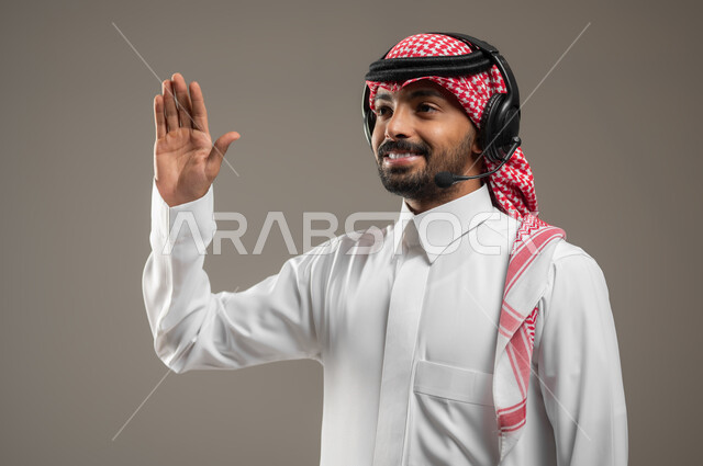 التواصل مع الأصدقاء والأقارب، استخدام تقنية الهيدفون، بورتريه مقرب لرجل عربي سعودي خليجي يرتدي الزي السعودي ويضع سماعه الرأس الصوتية، يشير بيده للاعلى، القاء التحيه والسلام بإيماءات السعاده، خلفيه رماديه، استخدام حساس، محتوى حساس