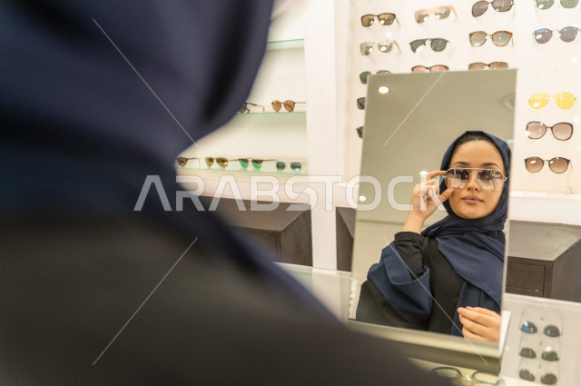 امرأة خليجية تقف امام المرآه  لاختيار نظارة شمسية مناسبة في محل نظارات , اختيار عصري للنظارات الشمسية