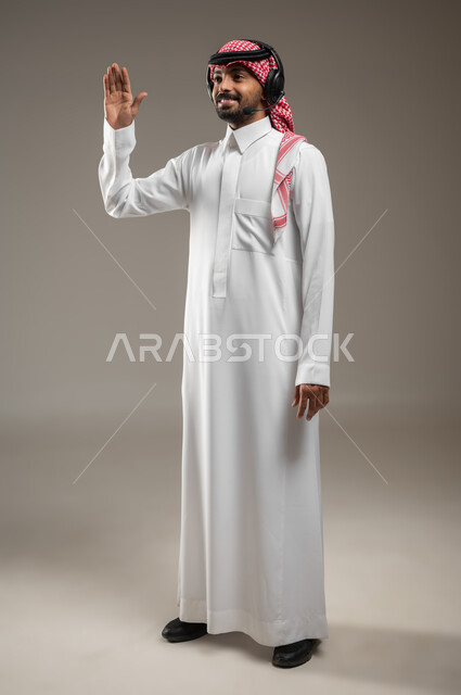 Communicating with friends and relatives, using headphone technology, a portrait of a Saudi Gulf Arab man wearing Saudi clothing and wearing an audio headset, pointing upward with his hand, saying hello and greetings with happy gestures, full-length body portrait, gray background, sensitive use, sensitive content
