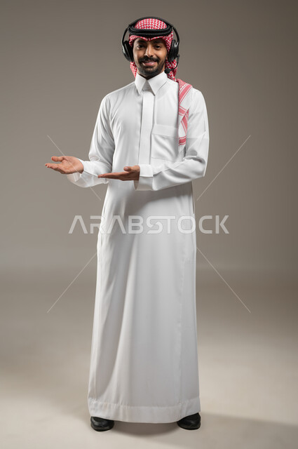 الاشارة الى شيء ما، استخدام الهيدفون للاستماع الى المقاطع الصوتية، بورتريه لرجل عربي خليجي سعودي يرتدي الزي السعودي يضع سماعة رأس يشير بابتسامه الى شئ ما، ايماءات وتعابير الترحيب، صوره الجسم بالطول الكامل، خلفيه رماديه، محتوى حساس، استخدام حساس