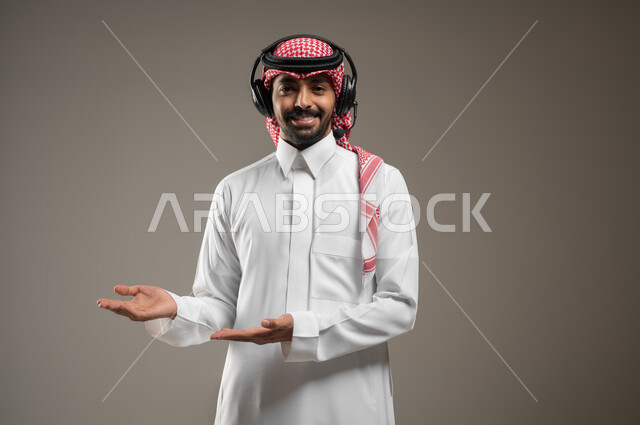 الاشارة الى شيء ما، استخدام الهيدفون للاستماع الى المقاطع الصوتية، بورتريه مقرب لرجل عربي خليجي سعودي يرتدي الزي السعودي يضع سماعة رأس يشير بابتسامه الى شئ ما، ايماءات وتعابير الترحيب، خلفيه رماديه، محتوى حساس، استخدام حساس