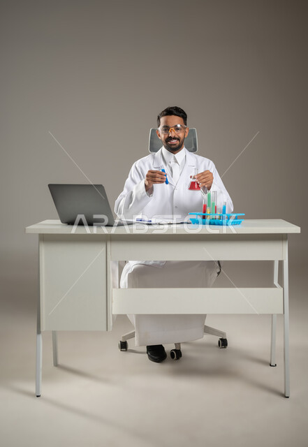 طبيب مخبري عربي خليجي سعودي يرتدي البالطو الابيض ،يجلس خلف مكتبه ينظر الى الكاميرا بأبتسامه، استخدام انابيب الاختبار لاجراء التجارب المخبريه والتفاعلات الكيميائية، مفهوم الكيمياء الحيوية، صوره المكتب بالطول الكامل، خلفيه رماديه، محتوى حساس، استخدام حساس