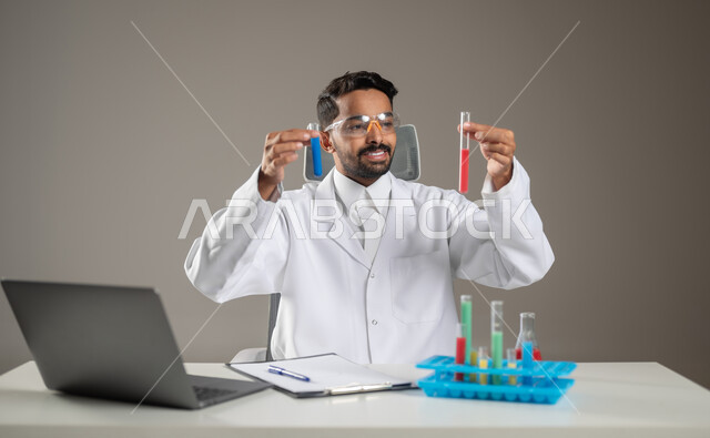 The importance of conducting chemical experiments and the development of teaching methods, gestures of precision, reasoning and concentration, a close-up portrait of a Saudi Arab Gulf laboratory doctor wearing medical protection glasses looking at laboratory tubes sitting behind his desk, scientific benefit from laboratory research, gray background, sensitive content