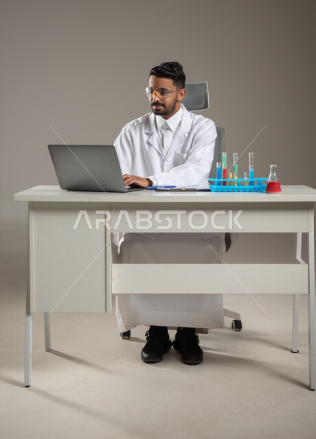 طبيب مخبري عربي خليجي سعودي يعمل من خلال جهازه المحمول في البحث والتطوير, الجلوس خلف المكتب، العمل عن بعد، تجارب وابحاث معمليه مخبريه، صوره المكتب بالطول الكامل، خلفيه رماديه، استخدام حساس، محتوى حساس