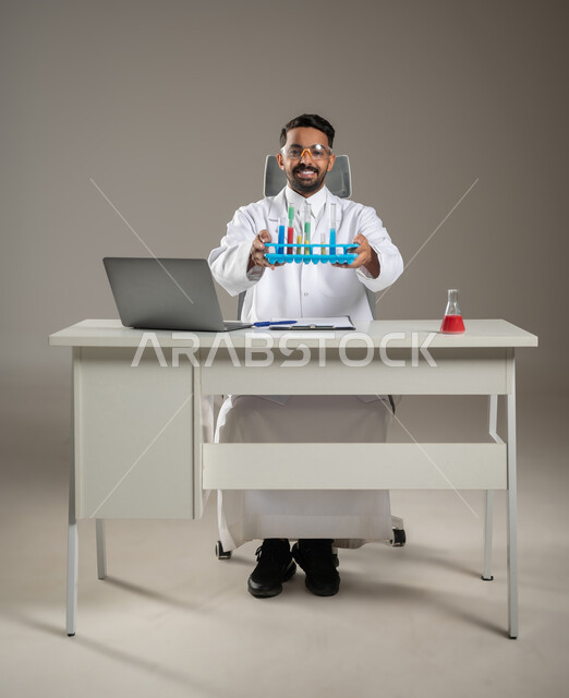 طبيب مخبري عربي خليجي سعودي مبتسم يقوم بعرض أنابيب إختبار التفاعلات الكيميائية جالسا خلف مكتبه، إجراء التحاليل الطبية ، تجارب و أبحاث علمية مخبرية ، مهن مستقبليه ، صوره المكتب بالطول الكامل، خلفية رماديه، محتوى حساس، استخدام حساس