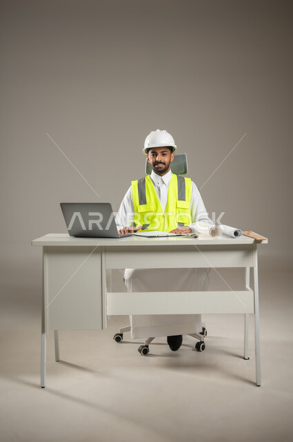 مهندس معماري سعودي خليجي يرتدي خوذه الحمايه وستره الوقايه، يتابع العمل على مخططات البناء ، يخطط لمشروع جديد ,ينظر الى الكاميرا بابتسامه، بورتريه لمهندس انشاءات خليجي بالثوب السعودي ، مخطط البناء، صوره المكتب بالطول الكامل ، خلفية رماديه، محتوى حساس
