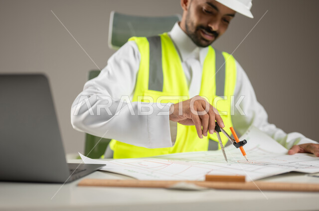 بورتريه مقرب لمهندس معماري سعودي خليجي يرتدي خوذه الحمايه البيضاء وستره الوقايه ، يتابع العمل على مخططات البناء ، يخطط لمشروع جديد , مهندس انشاءات خليجي بالثوب السعودي ، العمل خلف المكتب، خلفيه رماديه، محتوى حساس، استخدام حساس