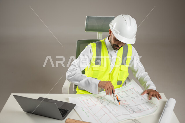 بورتريه مقرب لمهندس معماري سعودي خليجي يرتدي خوذه الحمايه البيضاء وستره الوقايه ، يتابع العمل على مخططات البناء ، يخطط لمشروع جديد , مهندس انشاءات خليجي بالثوب السعودي ، العمل خلف المكتب، خلفيه رماديه، محتوى حساس، استخدام حساس