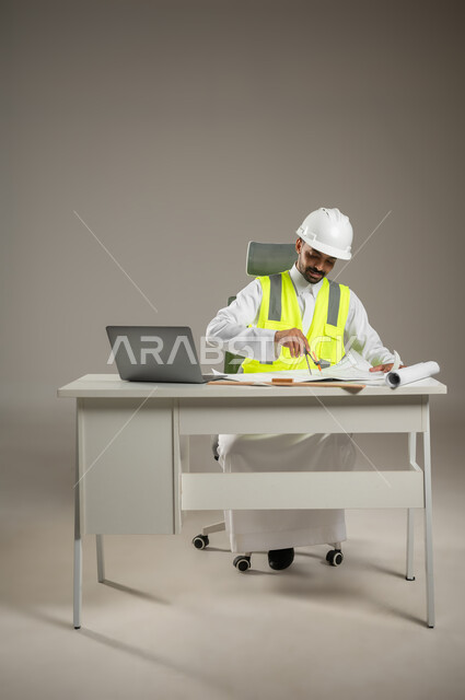 بورتريه لمهندس معماري سعودي خليجي يرتدي خوذه الحمايه البيضاء وستره الوقايه ، يتابع العمل على مخططات البناء ، يخطط لمشروع جديد , مهندس انشاءات خليجي بالثوب السعودي ، العمل خلف المكتب، صوره المكتب بالطول الكامل، خلفيه رماديه، محتوى حساس، استخدام حساس