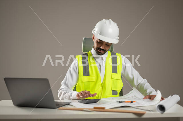 بورتريه مقرب لمهندس معماري سعودي خليجي يرتدي خوذه الحمايه البيضاء وستره الوقايه ، يتابع العمل على مخططات البناء ، يخطط لمشروع جديد , مهندس انشاءات خليجي بالثوب السعودي ، العمل خلف المكتب، خلفيه رماديه، محتوى حساس، استخدام حساس