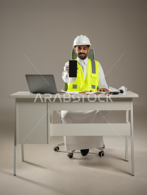 تطور ونمو القطاع الهندسي في السعودية، النظر إلى الكاميرا بإيماءات السرور، عرض شاشة سوداء فارغة، بورتريه لمهندس عربي سعودي خليجي يرتدي الخوذة وسترة الحماية،صوره المكتب بالطول الكامل، خلفية رمادية، استخدام حساس، محتوى حساس