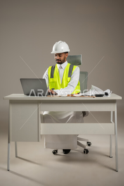 انجاز مهام العمل عن بعد، بورتريه  لمهندس عربي خليجي سعودي يرتدي الثوب السعودي والستره الوقائيه الخاصه بمهنه الهندسه، يرتدي خوذه الحمايه ويجلس خلف مكتبه، يؤدي عمله باستخدام الحاسوب، تصاميم ومخططات، صوره المكتب بالطول الكامل، خلفيه رماديه، محتوى حساس
