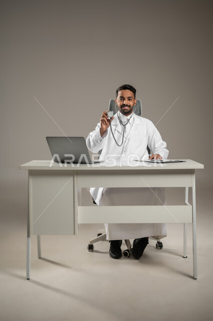 استخدام  الحاسوب في البحث والتطوير، بورتريه لطبيب عربي خليجي سعودي يرتدي المعطف الابيض ويضع سماعته الطبيه حول رقبته يجلس خلف مكتبه، يمسك بسماعته ويشير بها الى الكاميرا بأبتسامه، صوره المكتب بالطول الكامل، خلفيه رماديه، محتوى حساس، استخدام حساس