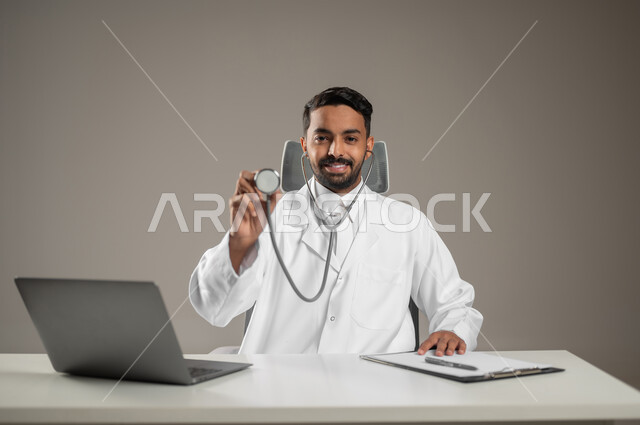 استخدام  الحاسوب في البحث والتطوير، بورتريه مقرب لطبيب عربي خليجي سعودي يرتدي المعطف الابيض ويضع سماعته الطبيه حول رقبته يجلس خلف مكتبه، يمسك بسماعته ويشير بها بأبتسامه الى الكاميرا، خلفيه رماديه، محتوى حساس، استخدام حساس