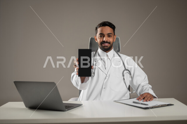 تقديم الخدمات للمرضى بكل كفاءه, اجراء الاستشارات الطبيه عن بعد, استخدام  الحاسوب في البحث والتطوير، بورتريه مقرب لطبيب عربي خليجي سعودي يرتدي المعطف الابيض ويضع سماعته حول رقبته، يمسك بجهازه المحمول يجلس خلف مكتبه، خلفيه رماديه، محتوى حساس، استخدام حساس