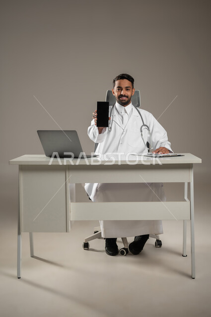 تقديم الخدمات للمرضى بكل كفاءه, اجراء الاستشارات الطبيه عن بعد, استخدام  الحاسوب في البحث والتطوير، بورتريه لطبيب عربي خليجي سعودي يرتدي المعطف الابيض يضع سماعته حول رقبته، يمسك بجهازه المحمول يجلس خلف مكتبه، صوره المكتب بالطول الكامل، خلفيه رماديه، محتوى حساس