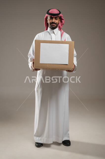 بورتريه لرجل عربي سعودي خليجي يرتدي الثوب والشماغ السعودي و يحمل بيده صندوق بورقه بيضاء فارغه، ينظر الى الكاميرا بأبتسامه، التخزين والتعبئه، صوره الجسم بالطول الكامل، خلفية رمادية، استخدام حساس، محتوى حساس