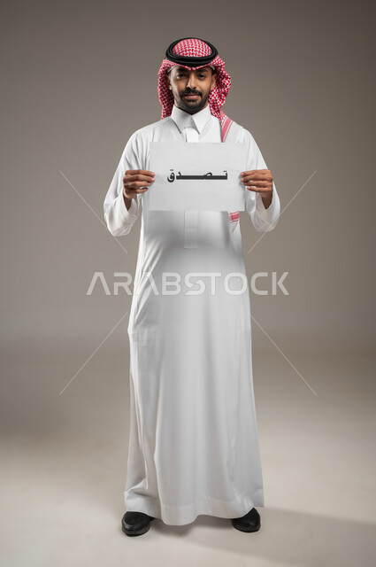 Islamic charitable works, humanitarian aid and donations, portrait of a Saudi Gulf Arab man wearing the Saudi thobe and shemagh and holding in his hand a paper with “Sadaqa” written on it, looking at the camera with pleased gestures, full-length body portrait, gray background, sensitive use, sensitive content
