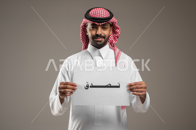 الأعمال الخيرية الإسلاميه، المساعدات والتبرعات الإنسانية، بورتريه مقرب لرجل عربي سعودي خليجي يرتدي الثوب والشماغ السعودي و يمسك بيده ورقه مكتوب بها تصدق، النظر إلى الكاميرا بإيماءات السرور، خلفية رمادية، استخدام حساس، محتوى حساس