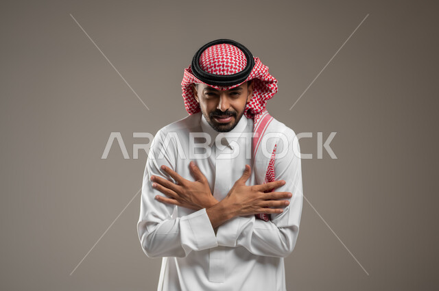الإحساس بالبرد الشديد والقارس في فصل الشتاء, بورتريه مقرب لشاب عربي خليجي سعودي  يرتدي الثوب والشماغ السعودي يقف بإيماءات تدل على الشعور بالبروده الشديده والالم، يضم يديه الى صدره، خلفية رمادية، محتوى حساس، استخدام حساس