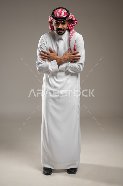 الإحساس بالبرد الشديد والقارس في فصل الشتاء, بورتريه لشاب عربي خليجي سعودي  يرتدي الثوب والشماغ السعودي يقف بإيماءات تدل على الشعور بالبروده الشديده والالم، يضم يديه الى صدره، صوره الجسم بالطول الكامل، خلفية رمادية، محتوى حساس، استخدام حساس