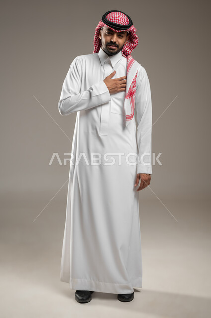 Gestures of fatigue, exhaustion, and psychological pressures, angina pectoris, a medical condition, a portrait of a Saudi Gulf Arab young man wearing the shemagh and the Saudi thobe, placing his hand on his heart, feeling a bad state of tingling and pain in his heart, full-length body photo, gray background, sensitive use, sensitive content