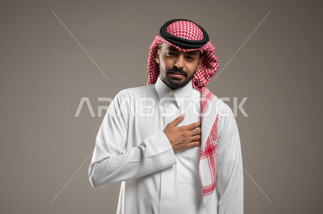 إيماءات التعب والإرهاق والضغوطات النفسية، الذبحه الصدريه حالة مرضية، بورتريه مقرب لشاب عربي سعودي خليجي يرتدي الشماغ و الثوب السعودي يضع يده على قلبه يشعر بحالة سيئة من الوخز والألم في قلبه، خلفية رمادية، استخدام حساس، محتوى حساس