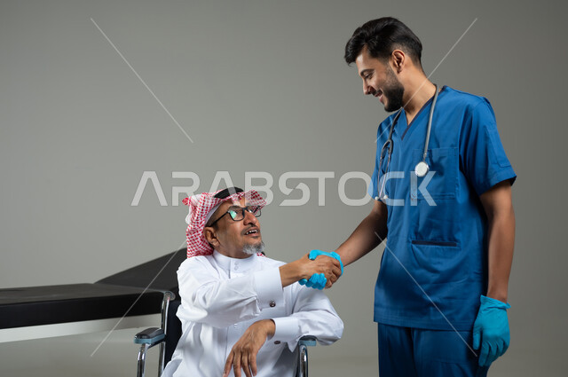 تقديم الاستشارة والخدمات الطبية لمرضى كبار السن في المستشفى، بورتريه لرجل عربي خليجي سعودي يرتدي الزي التقليدي والشماغ يجلس على الكرسي المتحرك يصافح الممرض ، شكر وتقدير الطبيب لجهوده ورعايته الصحية ، خلفية رمادية، محتوى حساس ، استخدام حساس