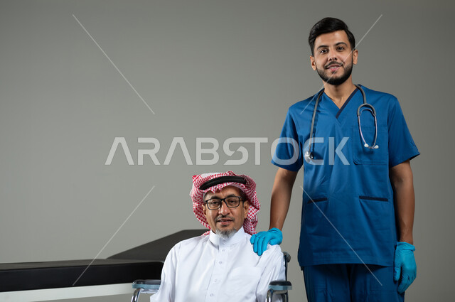 النظر إلى الكاميرا بإيماءات السرور، الاهتمام بصحة المرضى، بورتريه مقرب لممرض اسعافات اولية يرتدي اليونيفورم يضع يديه على كتف رجل عربي خليجي سعودي يرتدي الزي التقليدي والشماغ يجلس على الكرسي المتحرك ، خلفية رمادية، محتوى حساس ، استخدام حساس