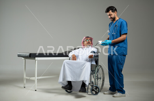 استخدام جهاز الضغط الالكتروني، بورتريه لممرض يرتدي اليونيفورم يقوم بقياس ضغط الدم لمريض عربي خليجي سعودي يرتدي الزي التقليدي والشماغ السعودي يجلس على كرسي متحرك، صورة الجسم بالطول الكامل، خلفية رمادية، محتوى حساس، استخدام حساس