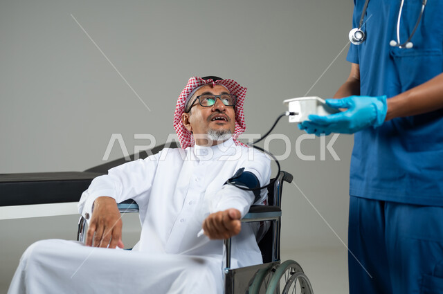 متابعة دورية لفحص ضغط الدم، التشخيص السريع والفحص الطبي والرعاية الصحية ، بورتريه مقرب لممرض يرتدي اليونيفورم يقوم بقياس ضغط الدم لمريض عربي خليجي سعودي يرتدي الزي التقليدي والشماغ السعودي يجلس على كرسي متحرك، خلفية رمادية، محتوى حساس، استخدام حساس