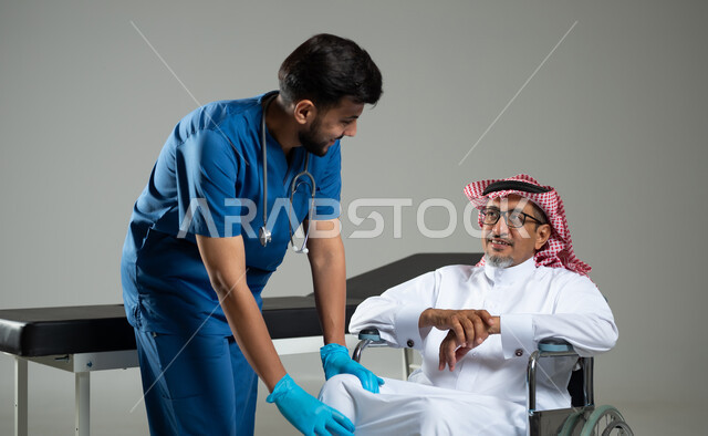 مفهوم الطب والرعاية الصحية، تقديم الخدمه والاحتياجات الخاصة بكل كفاءة، بورتريه مقرب لممرض عربي خليجي يرتدي اليونيفورم يقوم بفحص مفاصل مريض سعودي مقعد يرتدي الثوب التقليدي يجلس على الكرسي المتحرك، خلفية رمادية ، محتوى حساس، استخدام حساس .