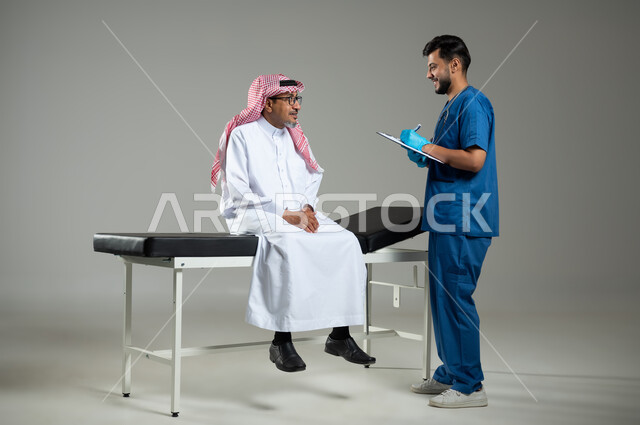 الوقوف باستقامة وتقديم الخدمات الصحية والطبية، بورتريه لطبيب عربي خليجي سعودي يرتدي اليونيفورم يقوم بتدوين المعلومات العامة لمريض مسن يرتدي الثوب التقليدي والشماغ يجلس على سرير المرضى, الجسم بالطول الكامل، خلفية رمادية، محتوى حساس ، استخدام حساس