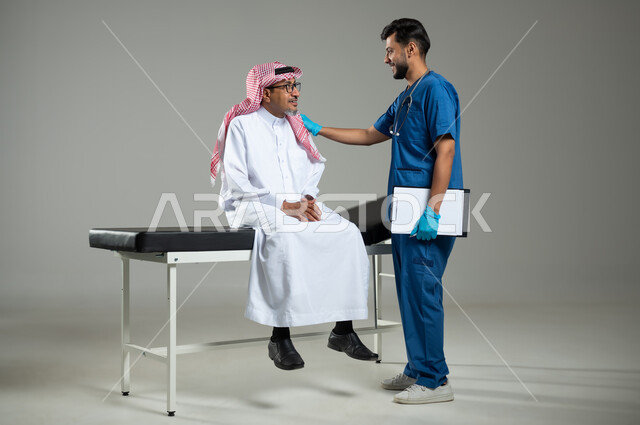 تقديم الخدمات الصحية , بورتريه لممرض يرتدي اليونيفورم يضع سماعة الأذن يقوم بالتأكد من سلامة اليد والكتف لرجل عربي خليجي سعودي يرتدي الثوب التقليدي والشماغ يجلس على سرير المرضى، مهن ووظائف طبية, الجسم بالطول الكامل، خلفية رمادية , محتوى حساس , استخدام حساس .