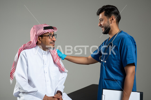 القيام بالرعاية الصحية , بورتريه مقرب لممرض يرتدي اليونيفورم يضع سماعة الأذن يقوم بالتأكد من سلامة اليد والكتف لرجل عربي خليجي سعودي يرتدي الثوب التقليدي والشماغ يجلس على سرير المرضى، مهن ووظائف طبية , خلفية رمادية , محتوى حساس , استخدام حساس .