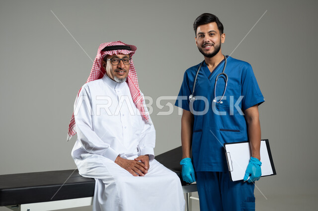 تقديم أفضل الخدمات الصحية لمرضى كبار السن في المستشفى, بورتريه مقرب لطبيب عربي خليجي سعودي يرتدي اليونيفورم يحمل بيده ملفات المرضى يفحص مريض عربي سعودي خليجي يرتدي الثوب التقليدي والشماغ يجلس على سرير المرضى، خلفية رمادية، محتوى حساس ، استخدام حساس