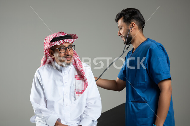 استخدام الأدوات الطبية الحديثة، اجراءات الفحص الطبي العامة لكبار السن، بورتريه مقرب لممرض عربي خليجي سعودي يقوم بتمرير جهاز سماعات الاذن على ظهر مريض مسن يرتدي الثوب التقليدي والشماغ يجلس على سرير المرضى، خلفية رمادية، محتوى حساس، استخدام حساس