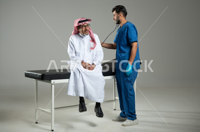 اجراءات الفحص الطبي العامة لكبار السن، بورتريه لممرض عربي خليجي سعودي يقوم بتمرير جهاز سماعات الاذن على ظهر مريض مسن يرتدي الثوب التقليدي والشماغ يجلس على سرير المرضى، استخدام الأدوات الطبية الحديثة، الطول الكامل للجسم، خلفية رمادية، محتوى حساس، استخدام حساس