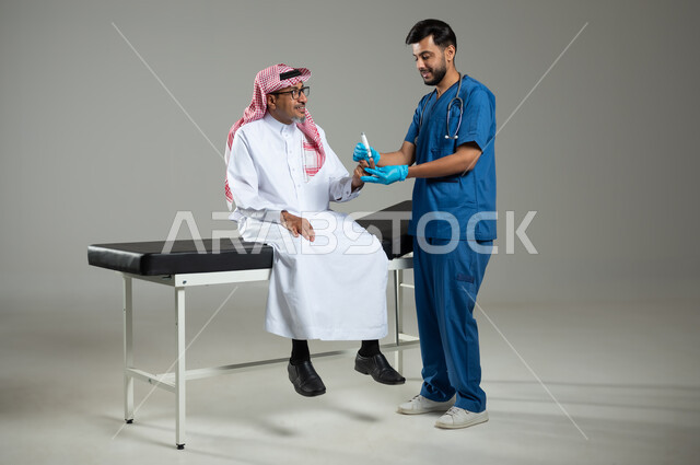 استخدام جهاز الفحص الالكتروني السكري، بورتريه لممرض عربي خليجي سعودي يرتدي اليونيفورم الطبي يقوم بفحص نسبة سكر الدم لمريض كبير بالسن يجلس على سرير المرضى, العمل في القطاع الصحي في السعودية، الجسم بالطول الكامل، خلفية رمادية, محتوى حساس، استخدام حساس